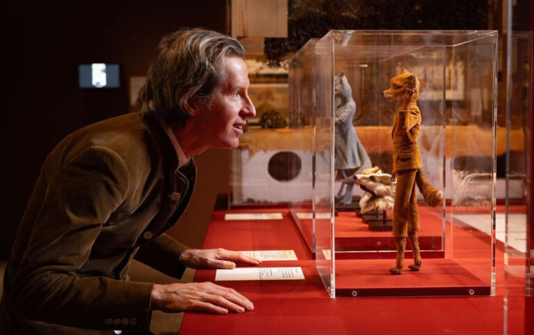 Wes Anderson beugt sich neugierig über eine Vitrine in einem Museum. Darin steht eine kleine Fuchspuppe in einem gelben Anzug. Die Szene ist warm beleuchtet, mit rotem Ausstellungstisch und weiteren Figuren in Glasvitrinen im Hintergrund.