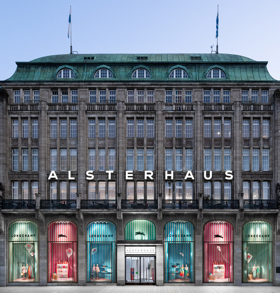 Das Alsterhaus als großes nordisches Haus gezeigt von der Fassade. Die unterste Etage zeigt sechs große Bögenschaufenster mit grünen, roten und blauen Designs für die Produkte von Longchamp.