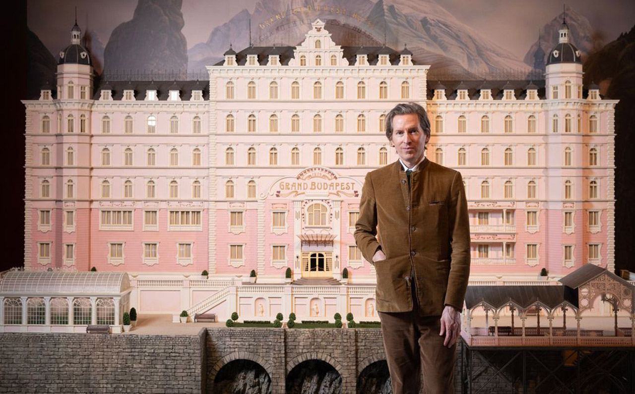 Wes Anderson, ein Mann mit braunem Anzug, steht vor einem großen, rosa Modell eines Hotels. Im Hintergrund sind aufgemalte Berge mit Schnee. 