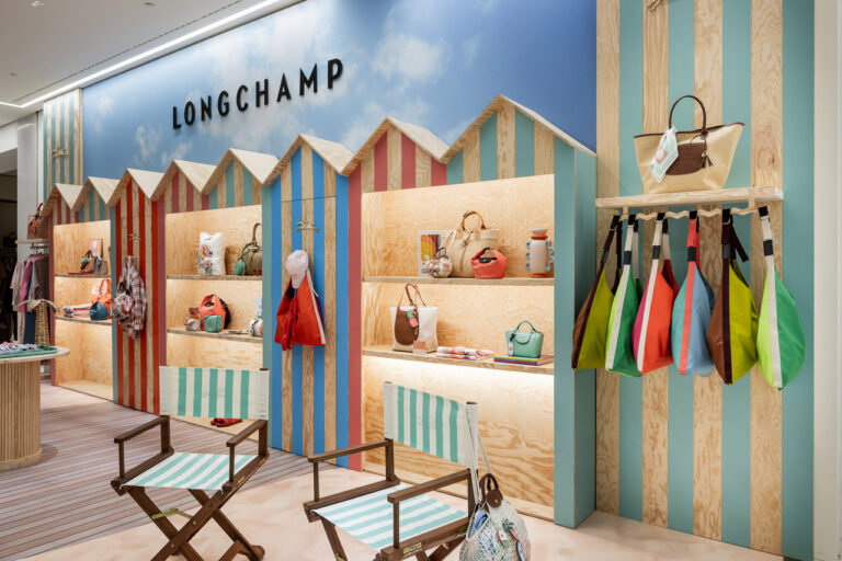 Verkaufsraum von Longchamp mit Strandoptik. Im Vordergrund stehen zwei grün weiß gestreifte Klappstühle. Ausgestellt sind bunte Taschen.