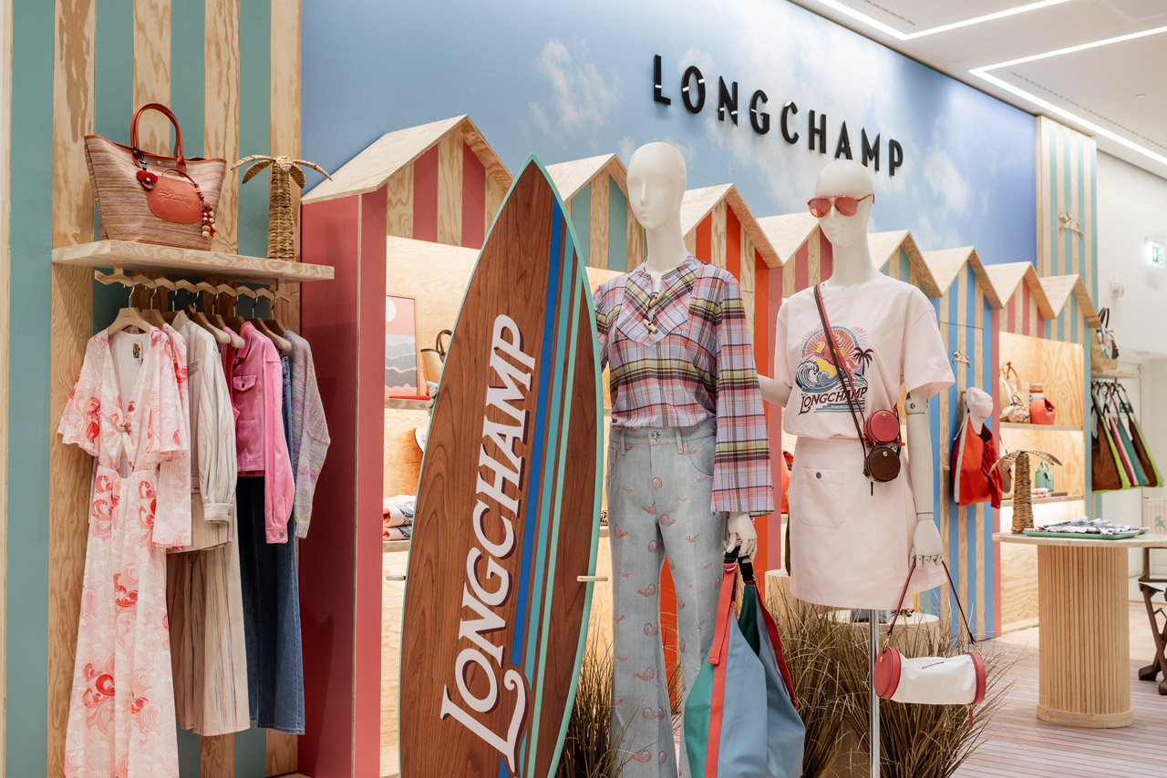 Ein bunter Austellungsraum im Strand Look mit hellen Farben von Longchamp. Im Vordergrund sind zwei Kleiderpuppen mit hellen Sommeroutfits. Die eine Puppe hält ein großes Longchamp Surfbrett aus Holz.