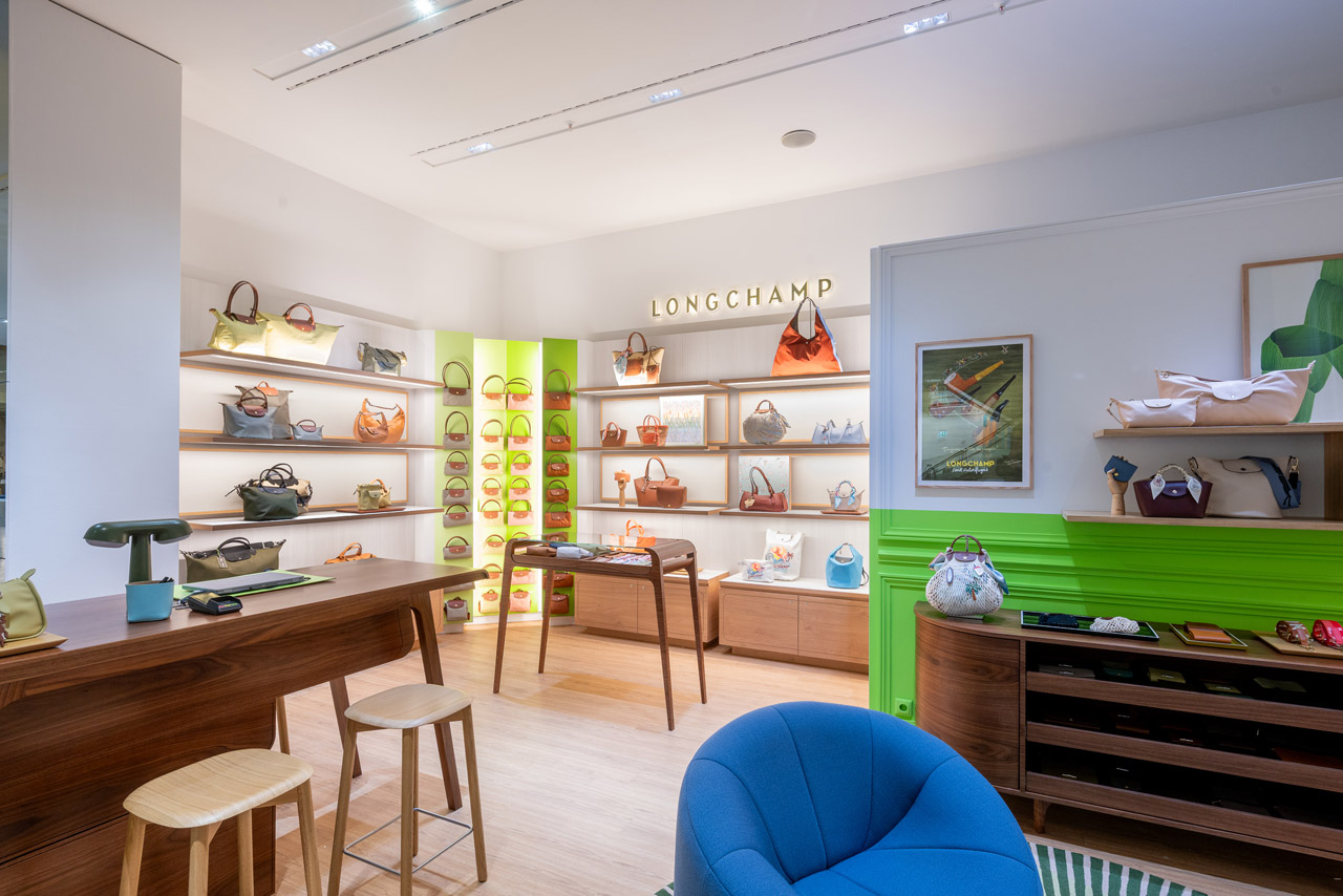 Ein offener, heller Ausstellungsraum von Longchamp. An den Wänden sind Taschen ausgestellt. Im Vordergrund ist ein Holztresen und ein gemütlicher blauer Sessel.