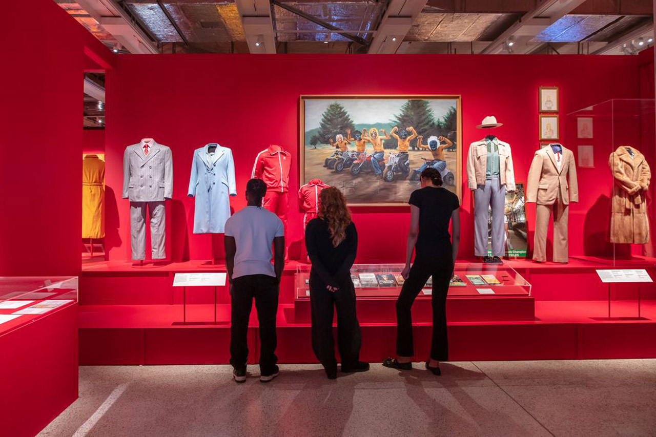 Drei Menschen stehen mit dem Rücken zum Betrachter und schauen sich Exponate in der Wes Anderson Ausstellung an. Die Kostüme, Bilder und Objekte sind vor einem leuchtend roten Hintergrund drapiert. Rechts im Bild ist ein langer, teuer aussehender Pelzmantel.